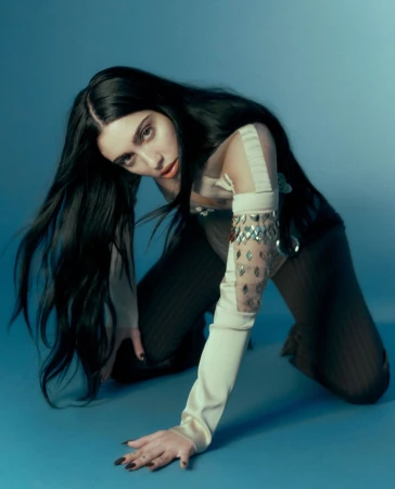 Brunette woman in tan and sheer clothing poses on her hands and knees against a gradient light-blue backdrop in a studio setting.