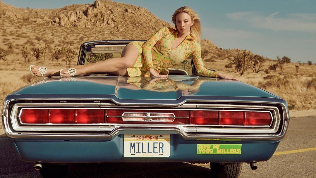 Blonde woman reclining on the hood of a classic blue convertible car in a desert landscape by Charlotte Wales.