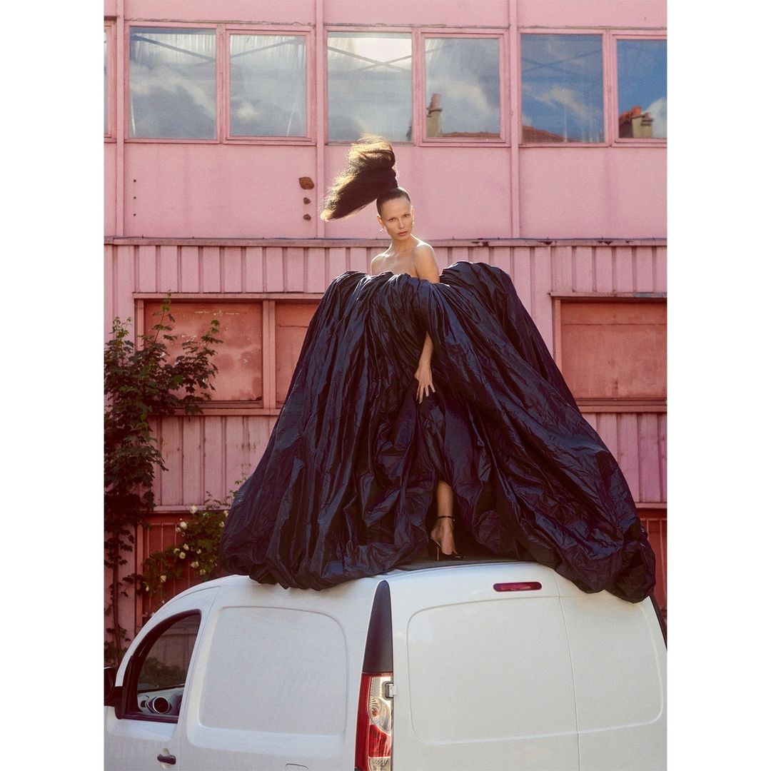 A woman in a large black dress stands on a white van, posing in front of a pink building, in a fashion editorial shot.