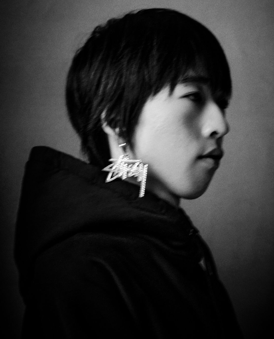 Monochrome portrait of a person with short black hair, wearing a hoodie and a large Stussy earring, gazing to the side in a studio.