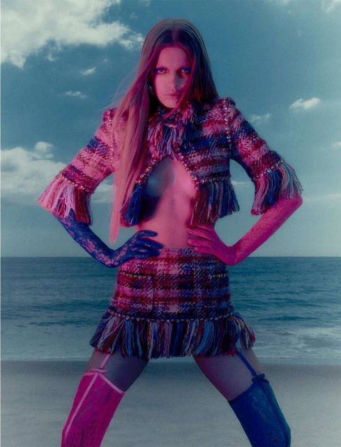 Fashion model poses on a beach wearing a tweed jacket, skirt, and color-blocked stocking in a surreal, vibrant fashion editorial.