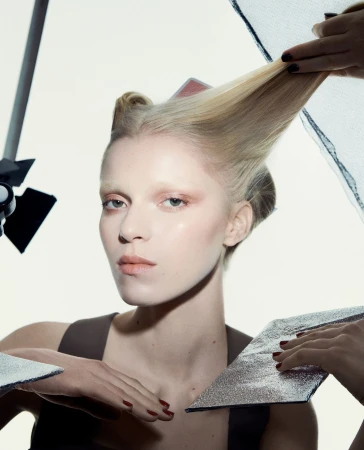 Blonde woman with stylized hair poses in a studio setting with lighting equipment and silver reflectors. Fashion and beauty shot.