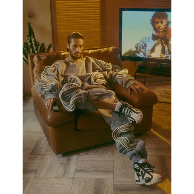 Adult male poses in a brown leather armchair, holding a remote, wearing an oversized gray sweatshirt and sweatpants set, with sneakers.