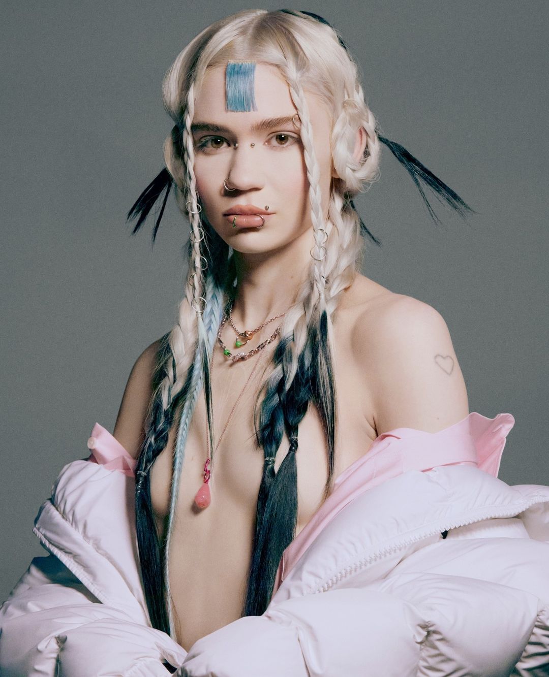 Portrait of woman with platinum blonde hair, braided with black and light blue strands, wearing a white puffer jacket in a gray studio.