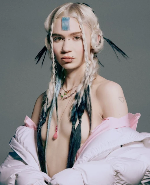 Portrait of woman with platinum blonde hair, braided with black and light blue strands, wearing a white puffer jacket in a gray studio.