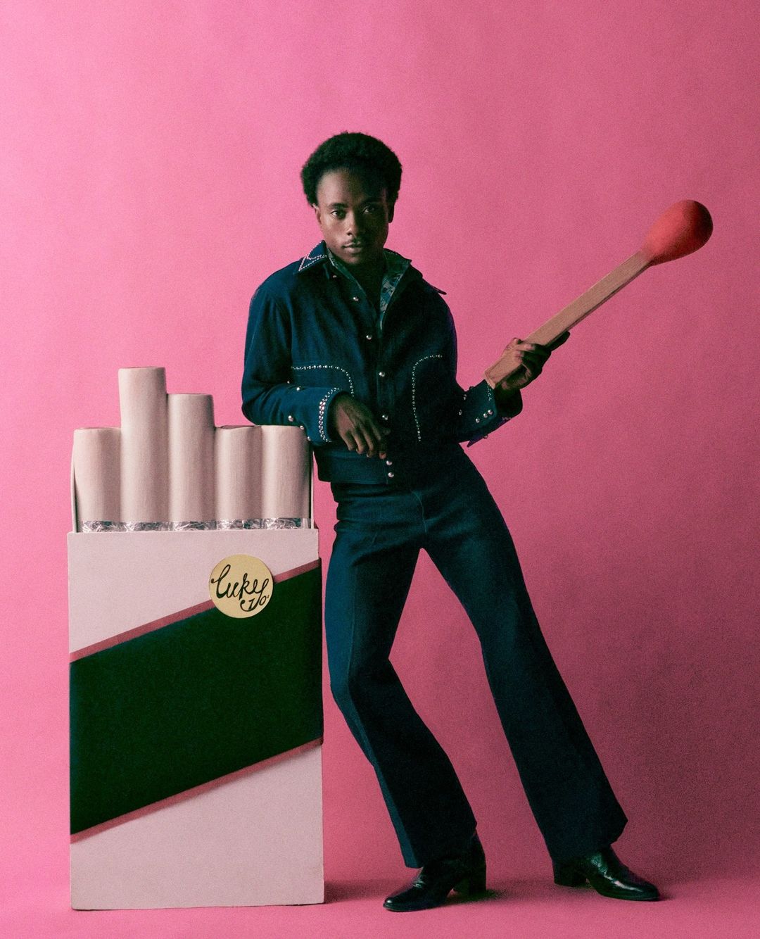 Man poses with a novelty cigarette pack and a match in a studio setting, wearing a dark denim suit against a pink backdrop.