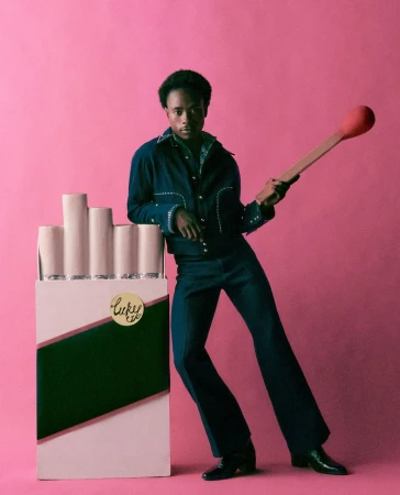 Man poses with a novelty cigarette pack and a match in a studio setting, wearing a dark denim suit against a pink backdrop.