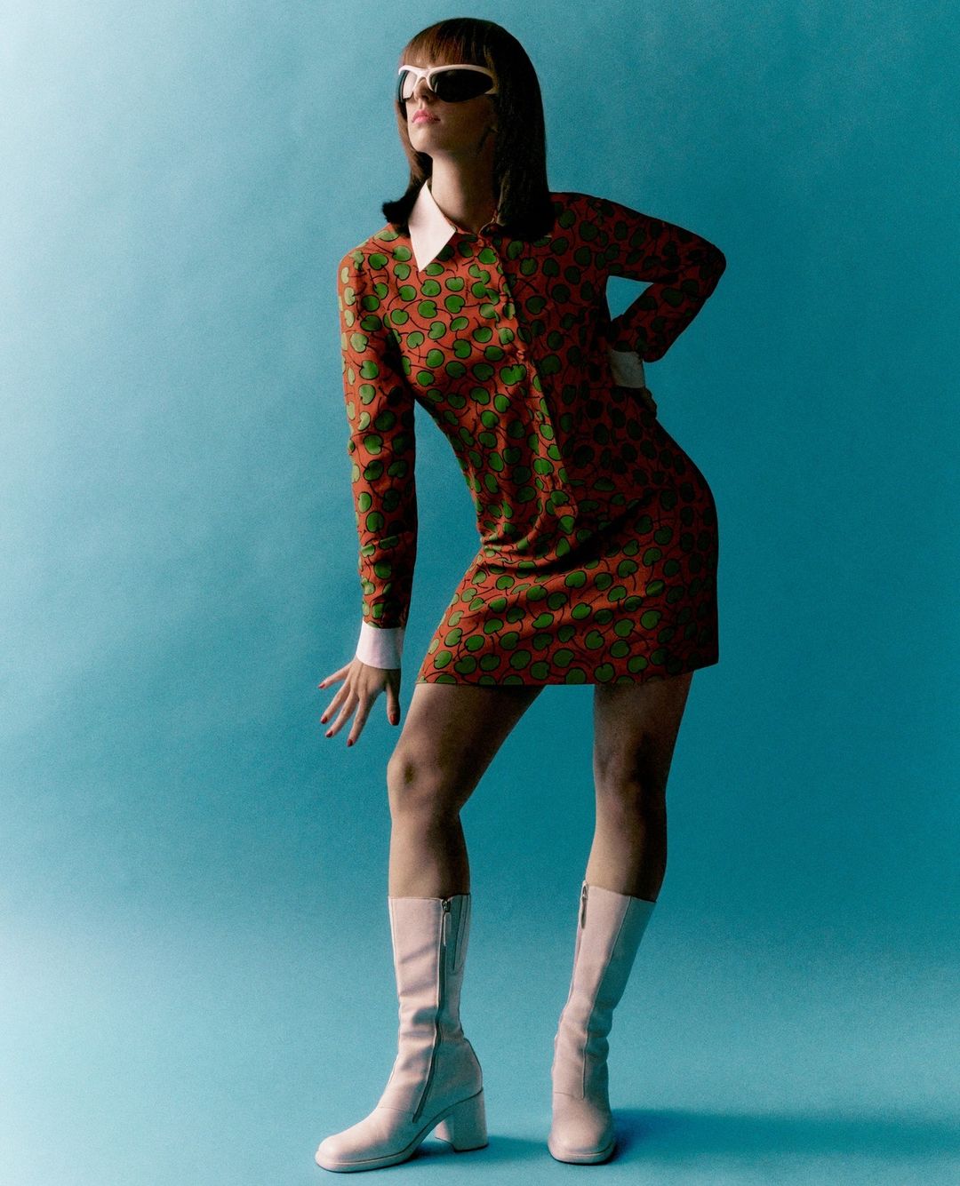 A stylish adult female poses in a retro-inspired orange and green mini dress with white accents and white boots against a blue background.
