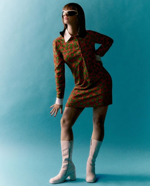 A stylish adult female poses in a retro-inspired orange and green mini dress with white accents and white boots against a blue background.