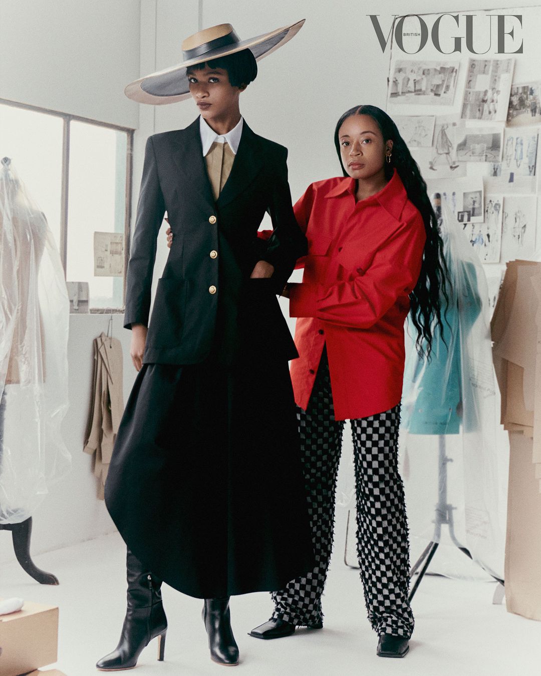 Two women pose in a bright studio; one wears a black suit and straw hat, the other a red shirt and checkered pants in a fashion editorial.