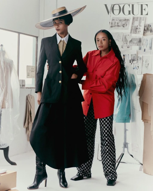 Two women pose in a bright studio; one wears a black suit and straw hat, the other a red shirt and checkered pants in a fashion editorial.