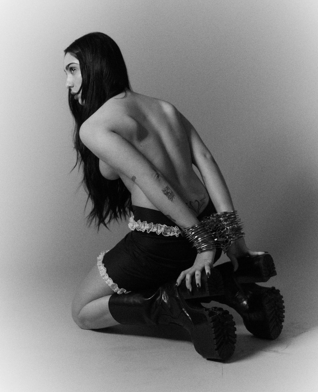 Monochromatic studio shot of a kneeling adult female with long dark hair wearing a skirt, bracelets, and platform boots.