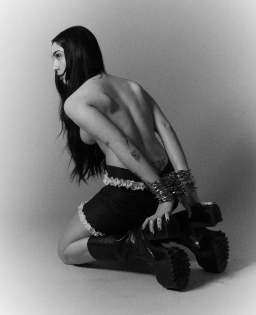 Monochromatic studio shot of a kneeling adult female with long dark hair wearing a skirt, bracelets, and platform boots.
