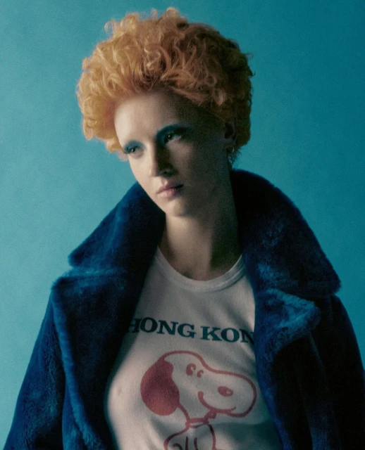 Young woman with curly orange hair poses in a white t-shirt and blue coat against a light blue backdrop in a fashion style photograph.