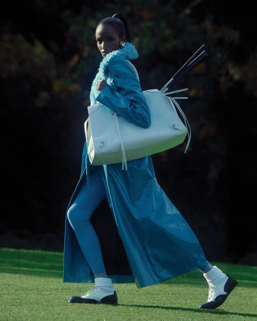 Fashion shot of a black female model walking in a turquoise outfit with a white purse against a dark foliage background on grass.