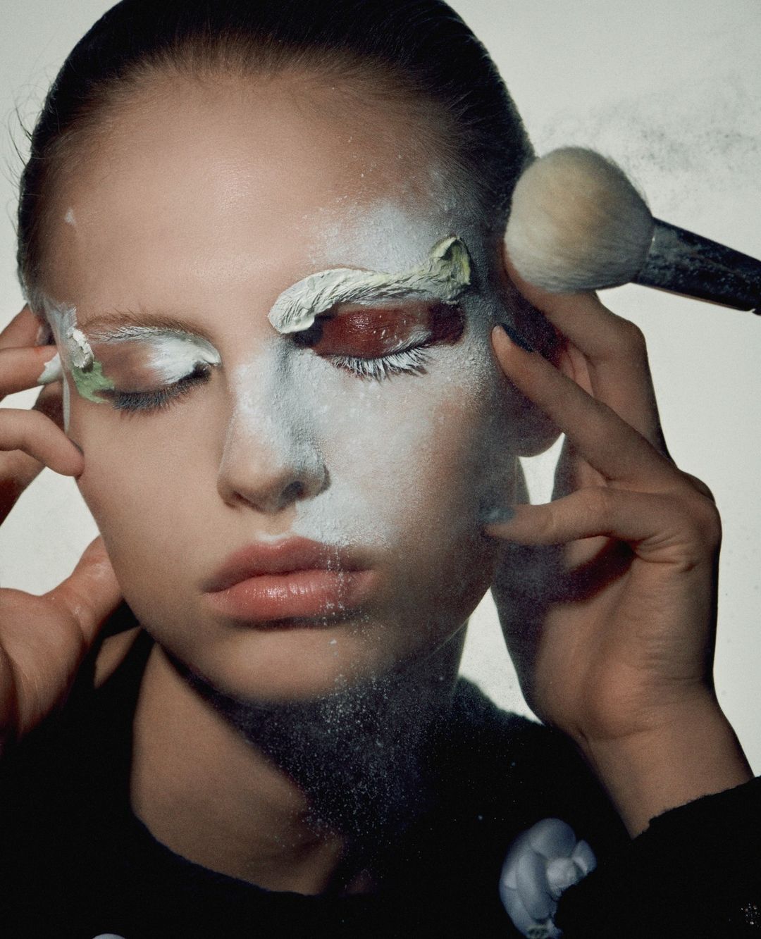 Closeup of a woman with white face paint and a makeup brush applying cosmetics near her eye in a bright, high-key lit studio.