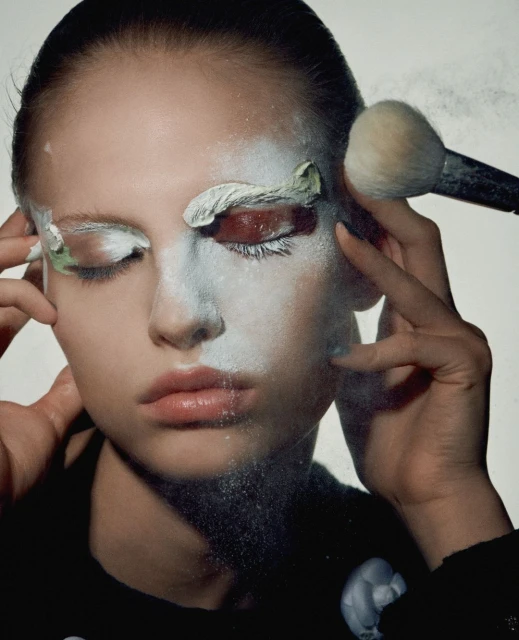 Closeup of a woman with white face paint and a makeup brush applying cosmetics near her eye in a bright, high-key lit studio.
