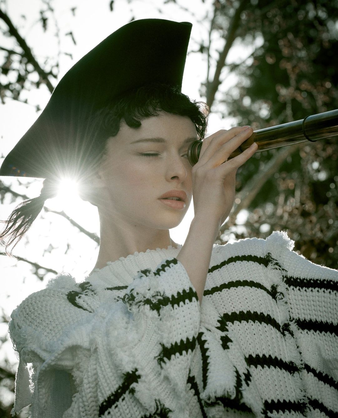 A woman in a whimsical hat peers through a telescope outside, wearing a white sweater with black stripes and a sunny outdoor setting.