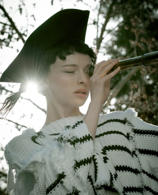 A woman in a whimsical hat peers through a telescope outside, wearing a white sweater with black stripes and a sunny outdoor setting.