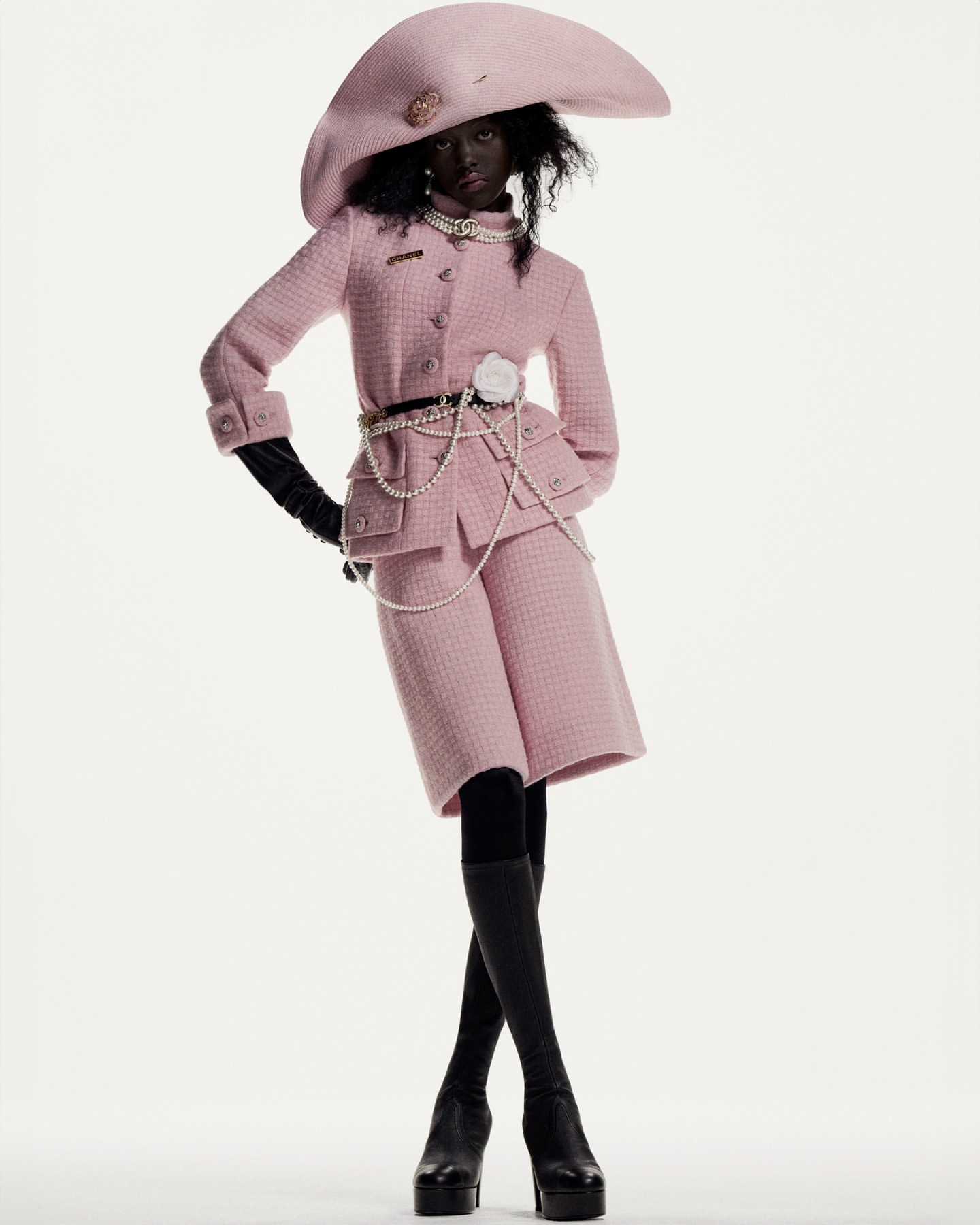 A dark-skinned woman posing in a pink tweed jacket, matching shorts, oversized hat, black gloves, black stockings, and platform heels.