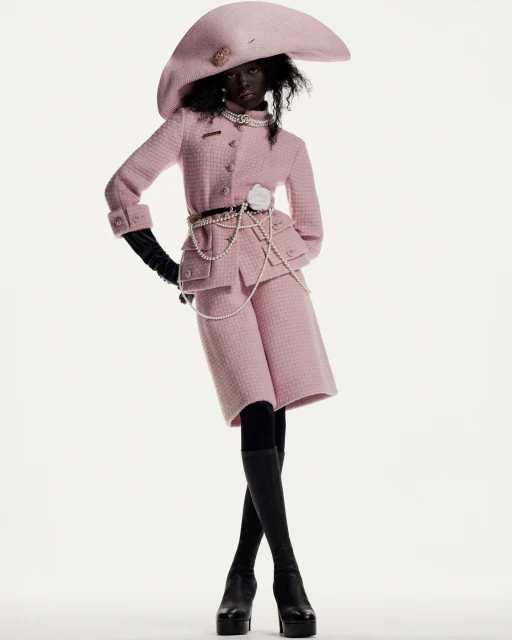 A dark-skinned woman posing in a pink tweed jacket, matching shorts, oversized hat, black gloves, black stockings, and platform heels.