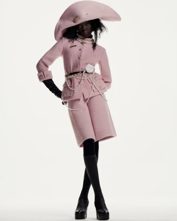 A dark-skinned woman posing in a pink tweed jacket, matching shorts, oversized hat, black gloves, black stockings, and platform heels.