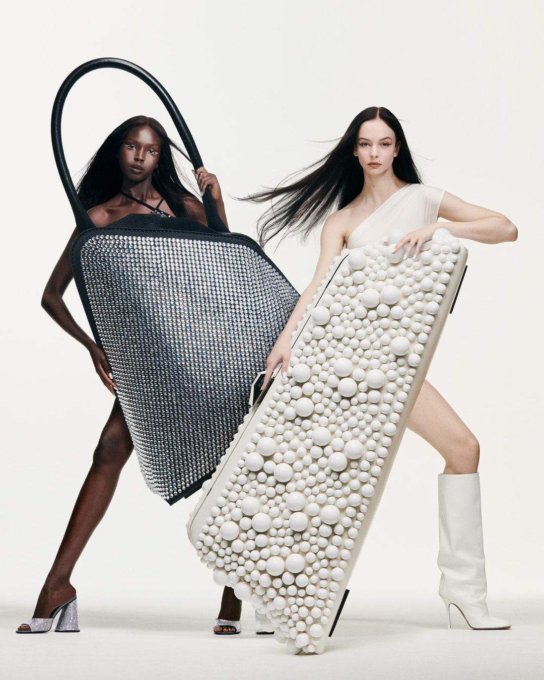 Two fashion models showcasing studded handbags in a studio. One model wears a black bag, the other a white bag, on a clean backdrop.