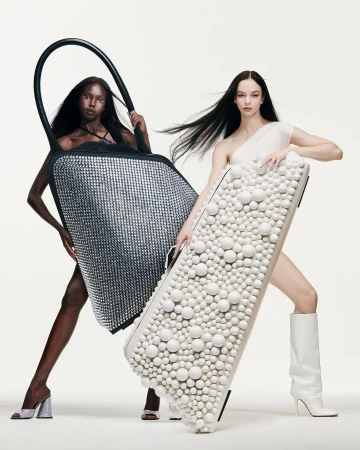 Two fashion models showcasing studded handbags in a studio. One model wears a black bag, the other a white bag, on a clean backdrop.