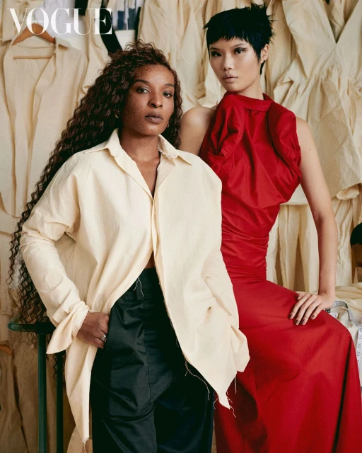 Editorial shot of two women posing in front of a wall of clothing; one in a cream shirt and black pants, the other in red dress. Vogue logo is visible.