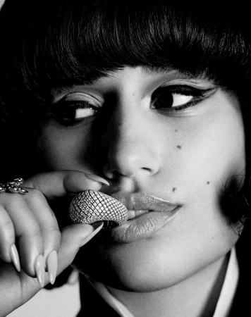 Close-up black and white portrait of a woman with heavy eye makeup holding a paved diamond ring near her lips.