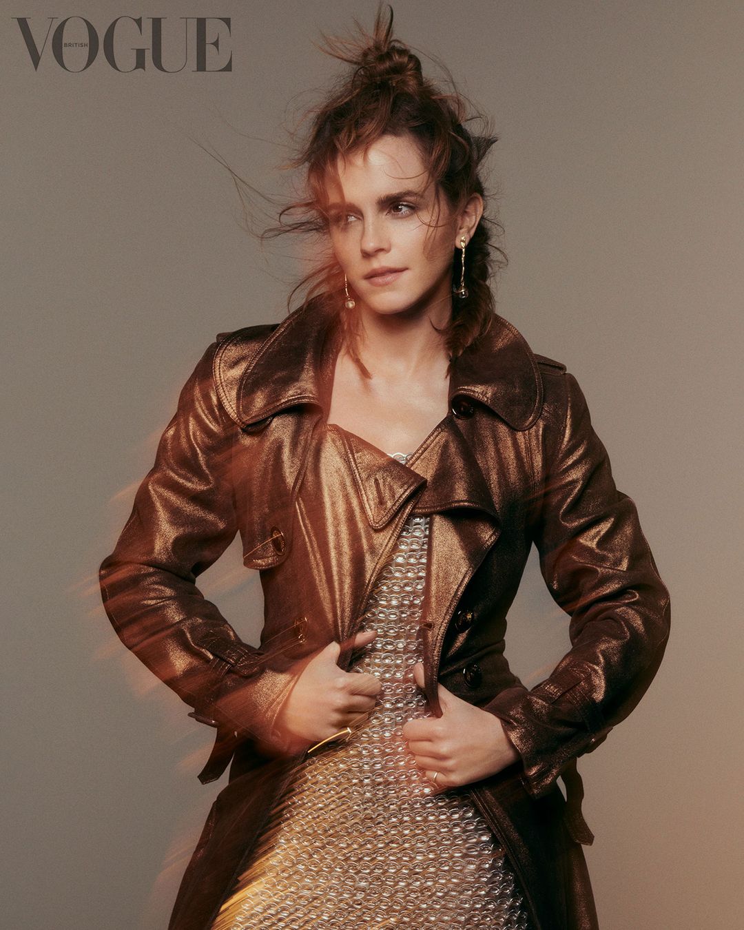 Emma Watson poses in a glossy brown trench coat and shimmery dress against a neutral backdrop for a Vogue magazine cover shoot.