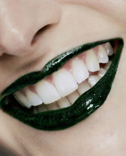Close-up of a smiling adult female with white teeth and shiny, dark green lipstick in a portrait-style composition indoors.
