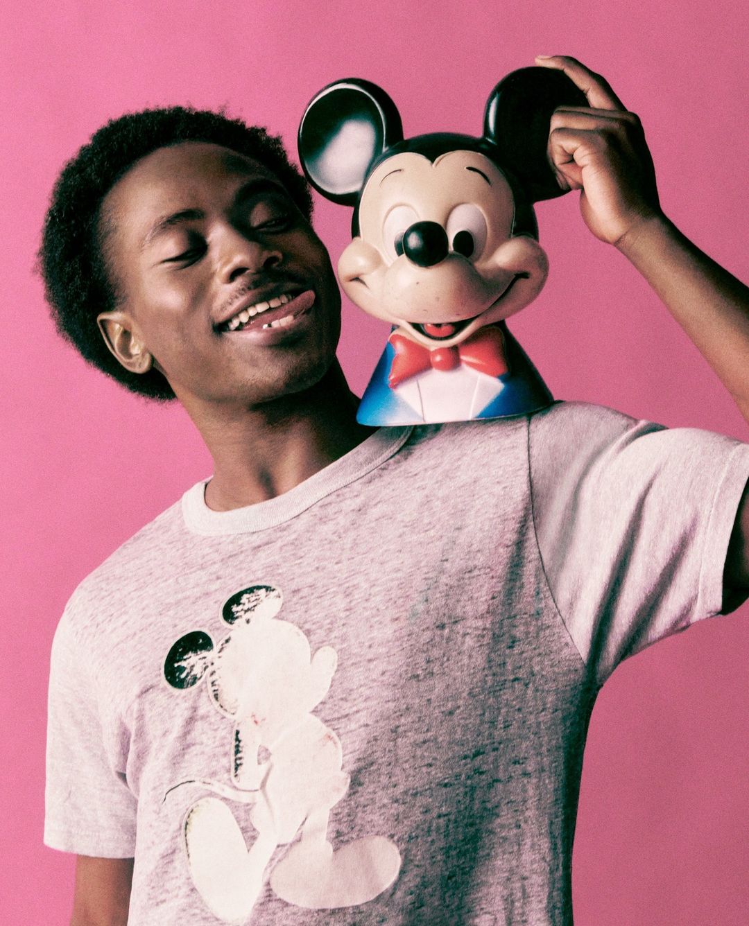 Smiling African-American man wearing a Mickey Mouse shirt and posing with a Mickey Mouse toy against a vibrant pink background.