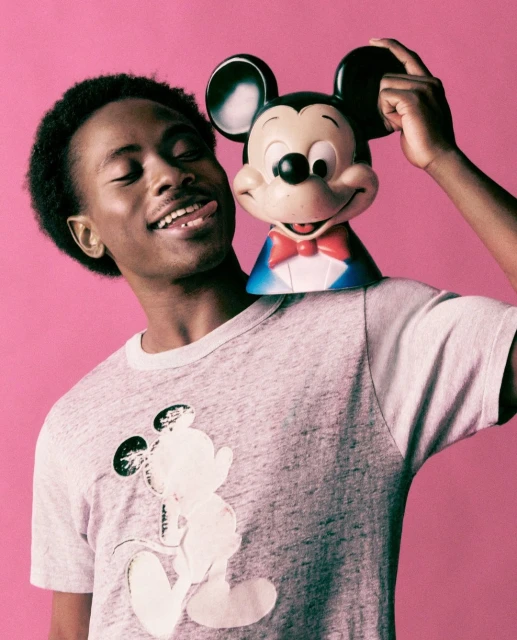 Smiling African-American man wearing a Mickey Mouse shirt and posing with a Mickey Mouse toy against a vibrant pink background.