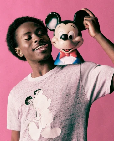 Smiling African-American man wearing a Mickey Mouse shirt and posing with a Mickey Mouse toy against a vibrant pink background.