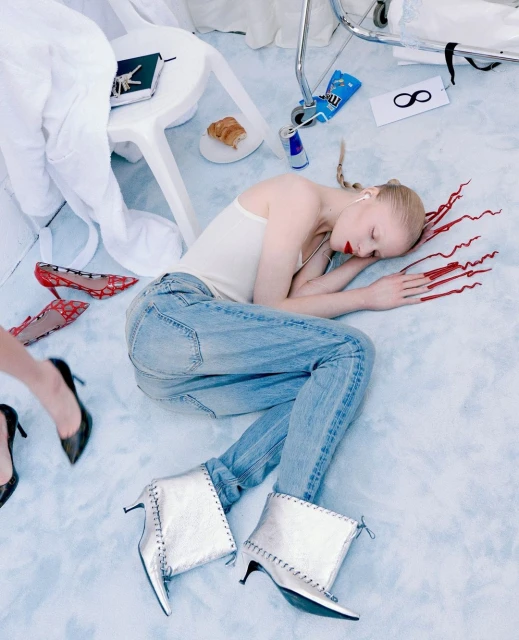 A woman reclines in silver boots and jeans on a light blue background, surrounded by shoes, food, and objects on a white surface.