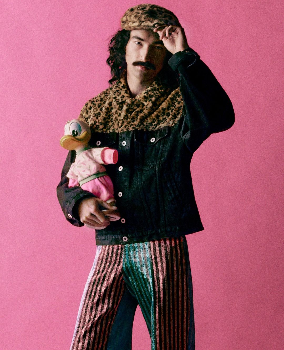 Person in leopard print hat and scarf holding a doll against pink background.  Striped pants add bold contrast to the unique style.