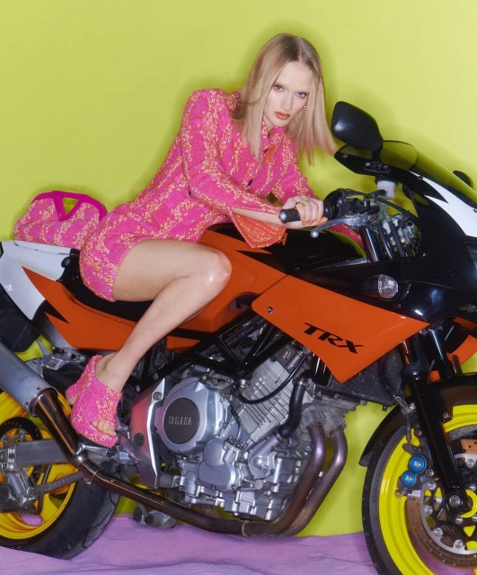 A fashionable blonde woman in a pink patterned dress and heels sits on a black and orange Yamaha TRX motorcycle against a yellow background.