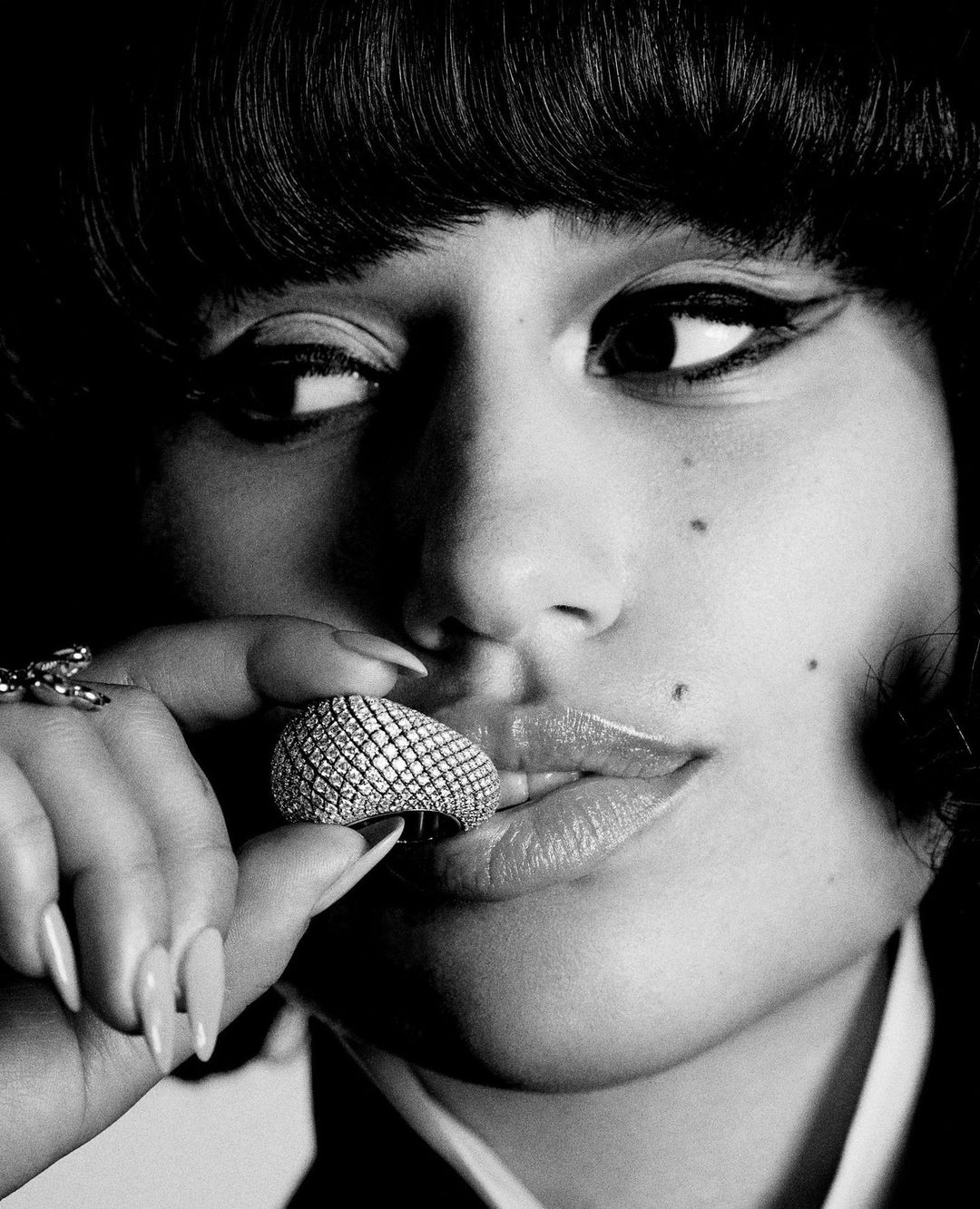 Close-up black and white portrait of a woman with winged eyeliner, holding a jeweled ring, showcasing beauty and jewelry.