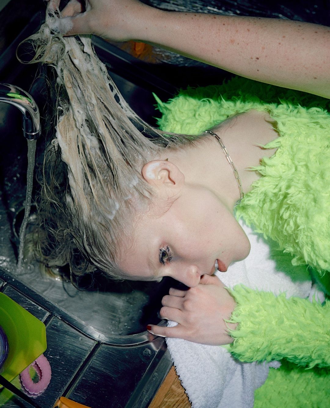 A fair-skinned adult woman with soapy blond hair leans over a stainless steel kitchen sink with a green sponge nearby; another person washes her hair.