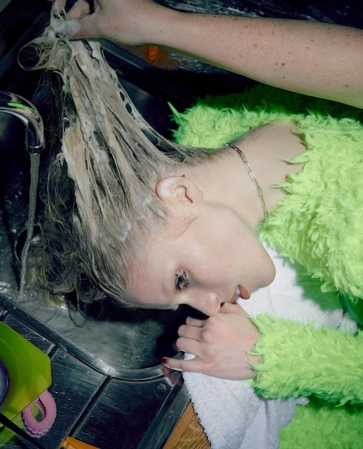 A fair-skinned adult woman with soapy blond hair leans over a stainless steel kitchen sink with a green sponge nearby; another person washes her hair.