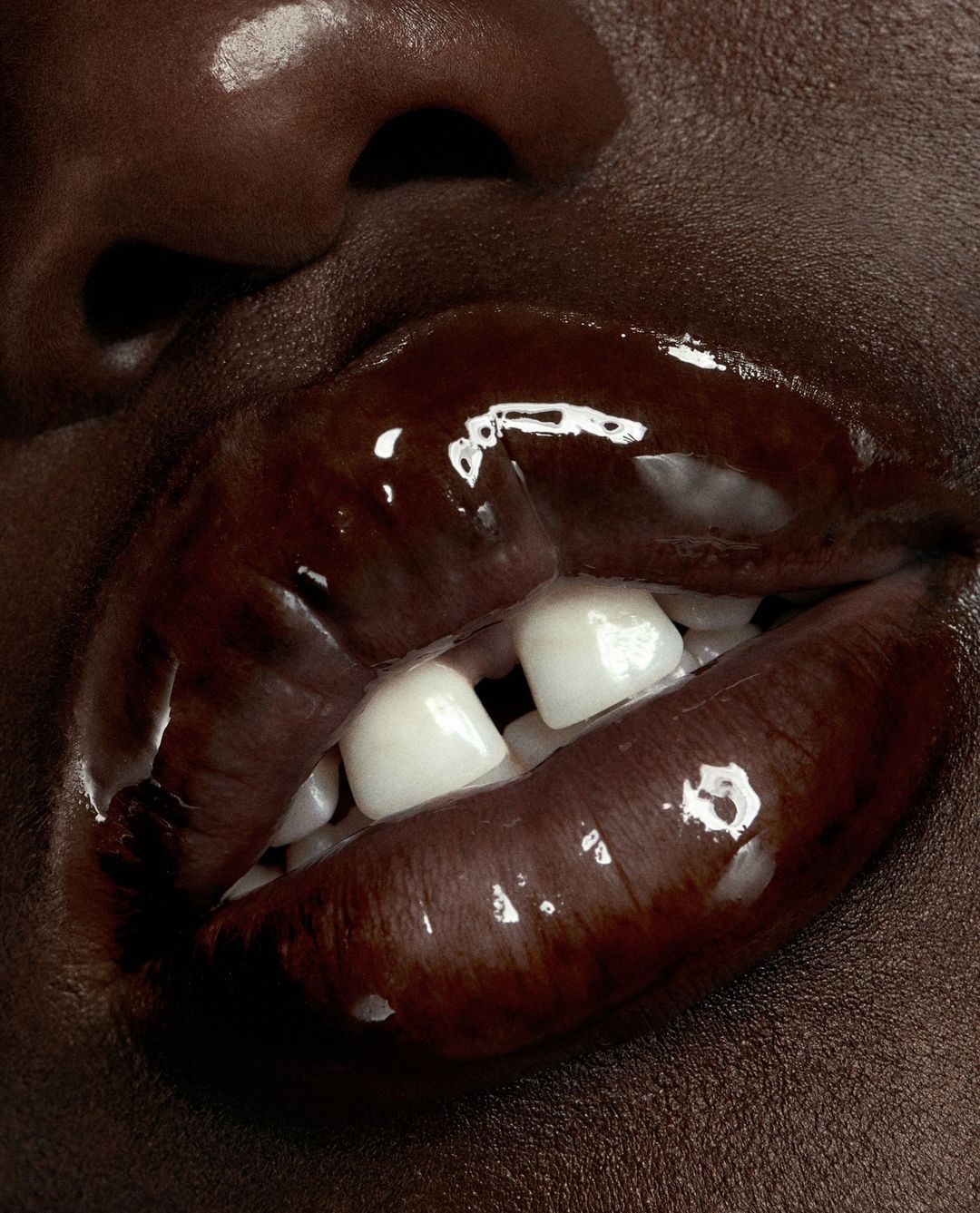 Close-up of an adult female's mouth with brown glossy lips, visible teeth, and dark skin, illuminated by studio lighting in an indoor setting.