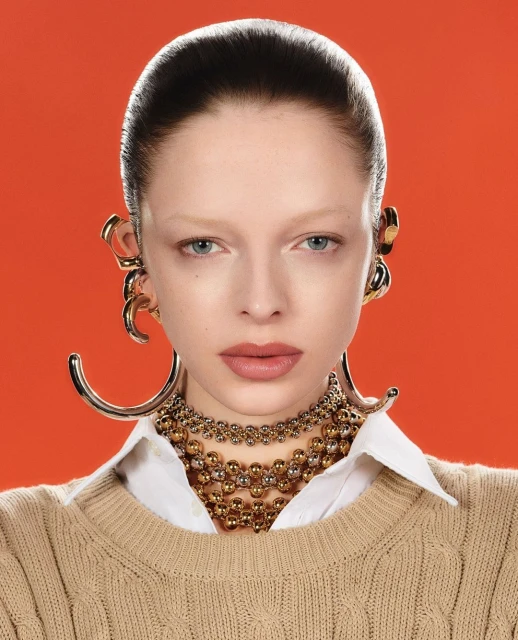 Close-up portrait of a woman with fair skin wearing gold ear cuffs, layered necklaces, a white button-down shirt and a knit sweater.