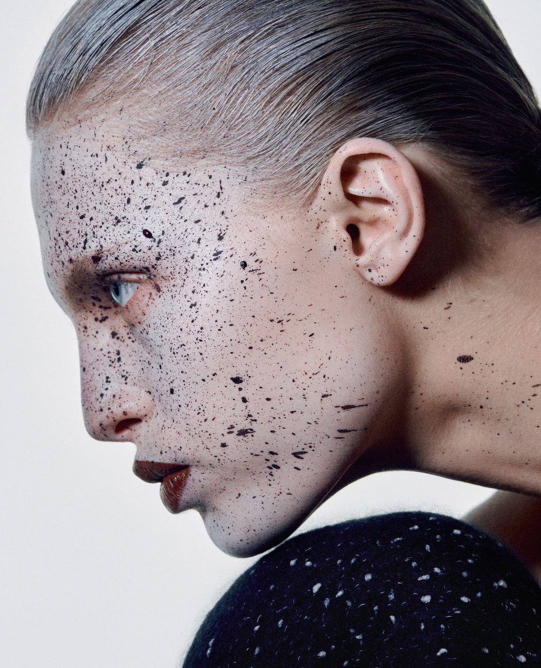 Close-up portrait of a woman with fair skin and dark hair, styled with speckled makeup, facing right on a seamless white background.