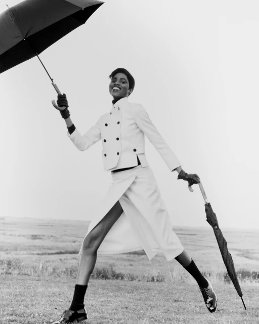 An adult female model in an all-white suit strides confidently in a black-and-white outdoor shot, holding two umbrellas in a windy field.
