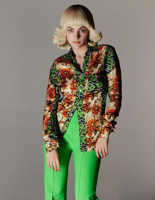 Adult woman in 1970s-inspired fashion posing in photo studio. Floral long-sleeve shirt, high-waisted bright green pants, and blonde bob wig.