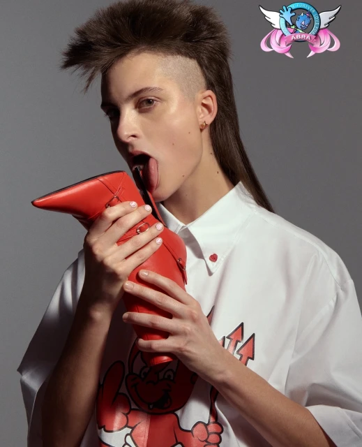 Stylish young woman poses with a red high-heeled boot in a fashion studio setting. She is wearing a white shirt with a devil graphic.