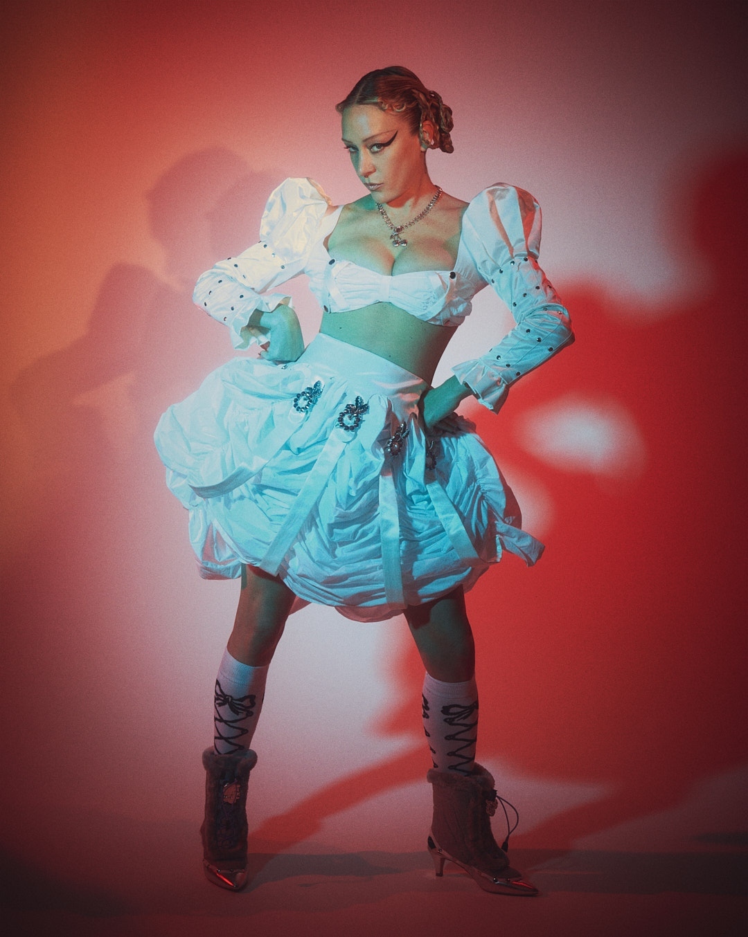 Woman in a white, frilly outfit poses in front of a gradient orange and blue background in a fashion-forward portrait.