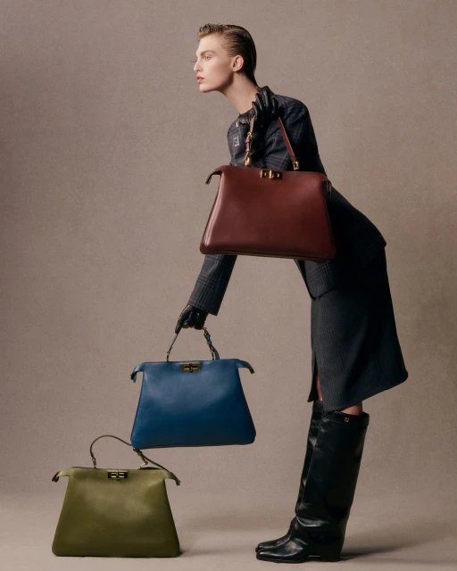 Fashion shot of a woman in a gray suit and black boots, posing with a collection of three colorful leather handbags in a studio.