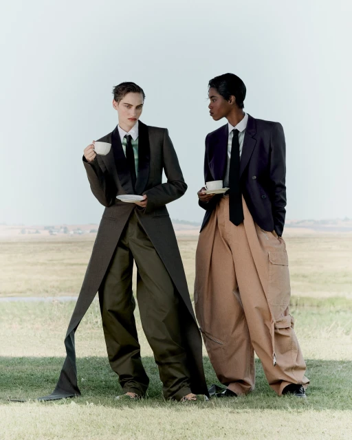 Two stylish adults outdoors in oversized suits, holding tea cups and saucers against a grassy field and pale sky background.